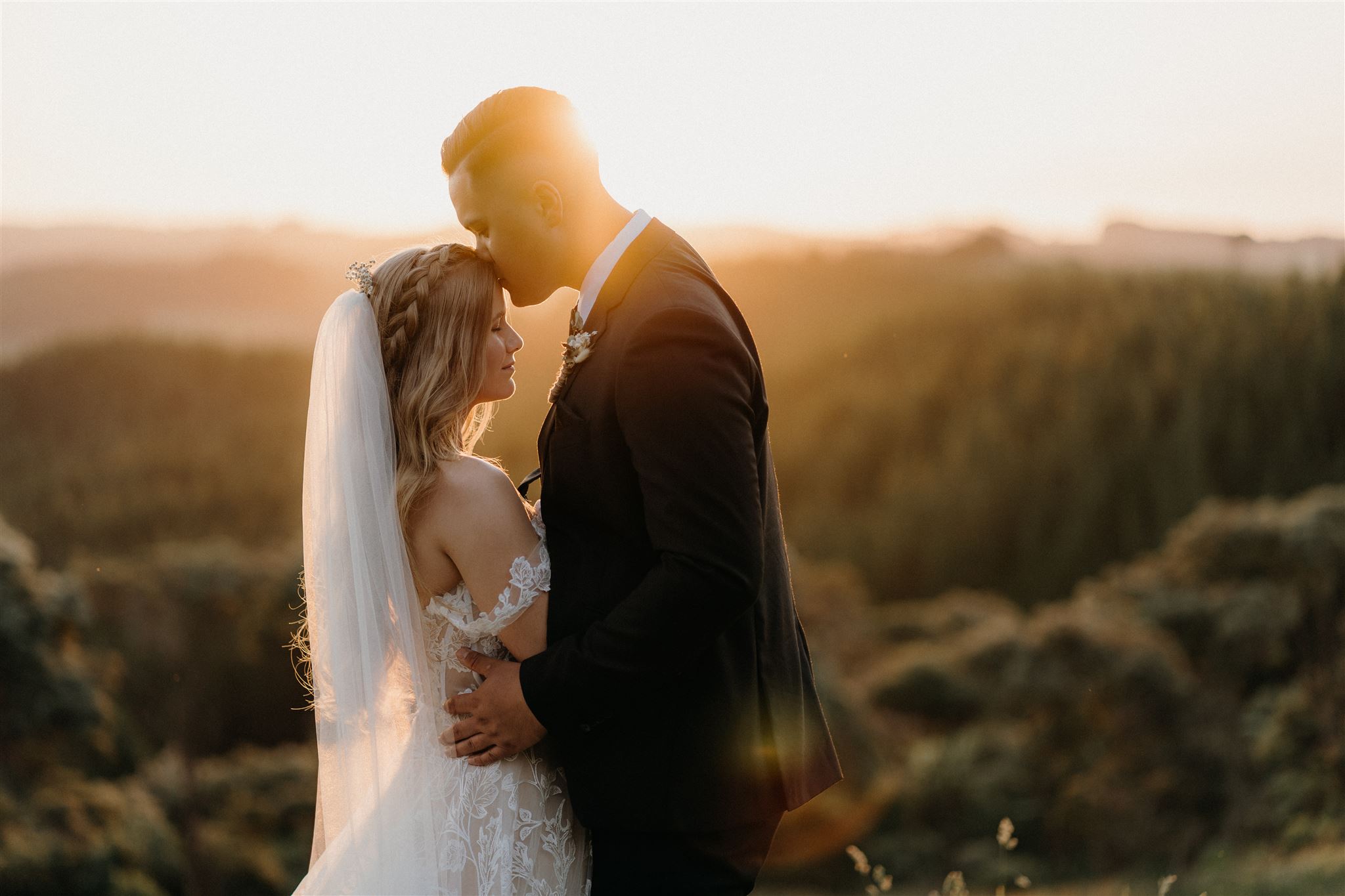 A groom kisses his new bride on the forehead during their wedding day photoshoot with Tashina Narelle Photo + Video while the sun sets behind them over the rolling hills.