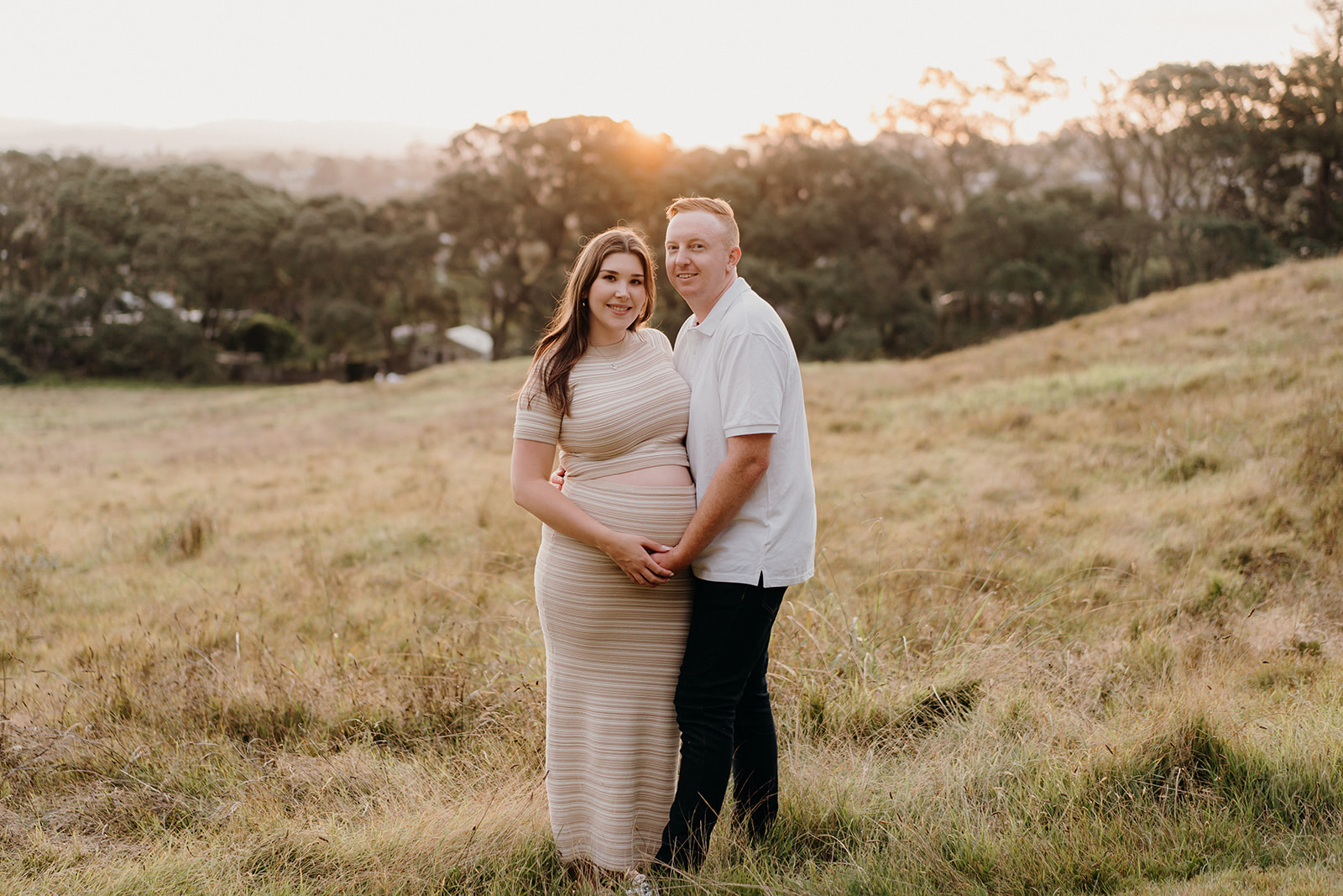 A pregnant woman and her partner stand and smile at the camera during their sunset maternity session with Tashina Narelle Photo + Video