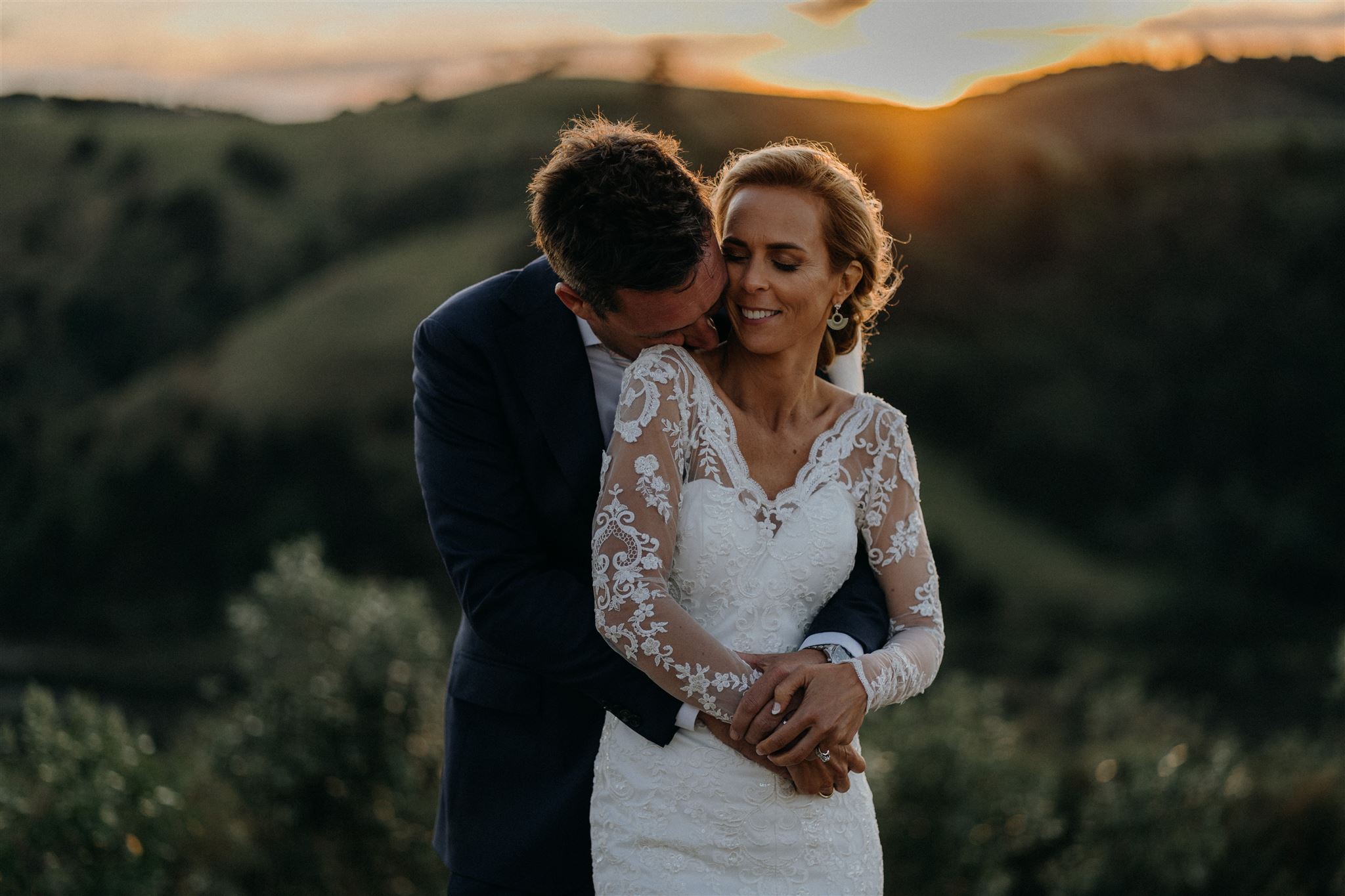 A Bride and Groom stand on top of the hill behind Poderi Crisci Wedding Venue Waiheke as the sun sets behind the hills they overlook during their photo session with Tashina Narelle. All loved up