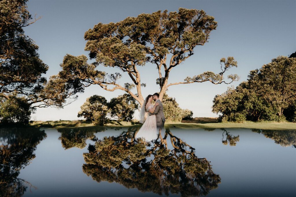 A photo of a bride and groom embracing during their portrait session after theri wedding at Wenderholm Regional Park, Auckland. They stand beneath a large Pohutukawa tree with the sky and tree reflected in the foreground using a creative reflection technique by Tashina Narelle 