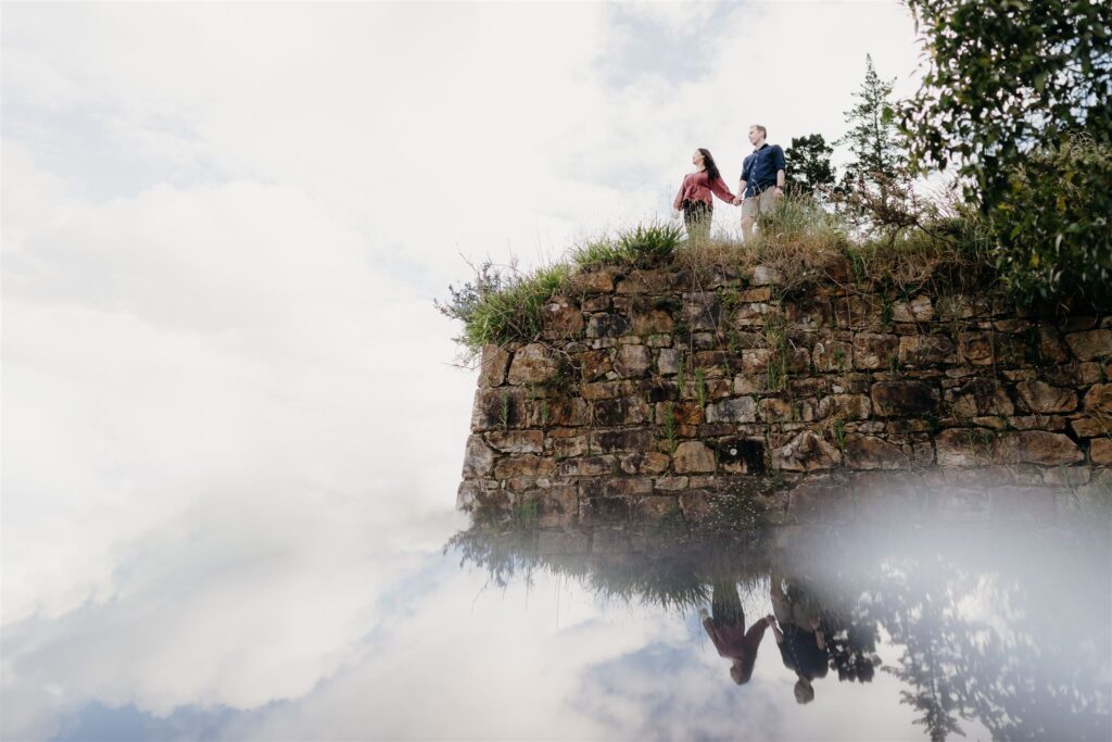 A couple stand on the ruins of an old retaining wall during their engagement photoshoot at Karangahke gorge.  The sky is reflected and they appear to be floating